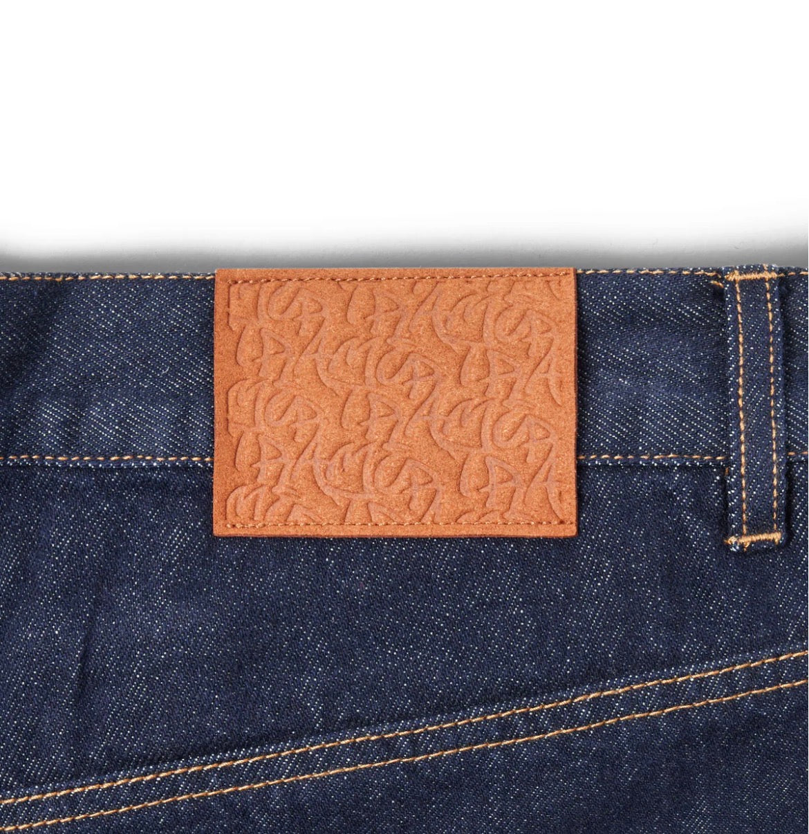 Close-up of a blue denim fabric with a brown leather label featuring an embossed pattern on a white background.